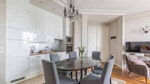 A modern kitchen with white cabinets, a round dining table with grey chairs, a chandelier, and a flat-screen TV in the living area. - Stunning & Bright, Sleeps 6, w/Big Balcony and Amazing Paris Views (Paris)
