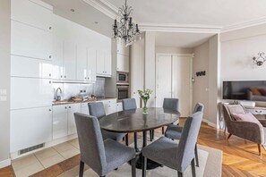 Una cocina moderna con armarios blancos, una mesa de comedor redonda con sillas grises, una araña y un televisor de pantalla plana en la zona de estar.