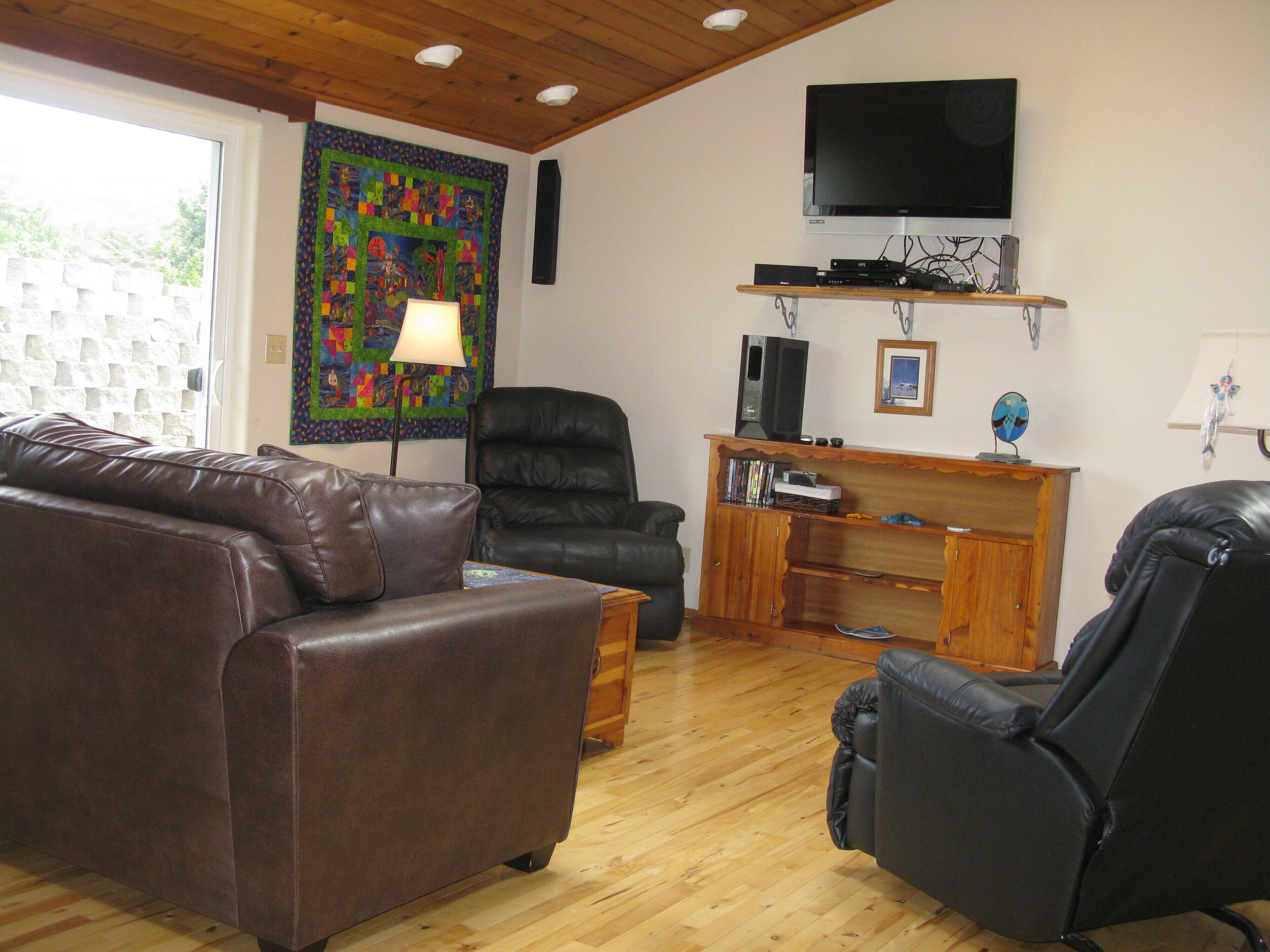 Una sala de estar con un sofá de cuero marrón, un sillón de cuero negro, un mueble de madera para entretenimiento con un televisor y un colcha colorida en la pared.