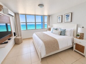 A hotel room with a large bed, a television, a window with a view of the ocean, and a small bedside table.
