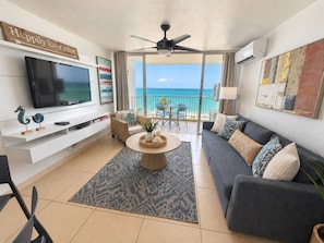 A living room with a flat-screen TV, a ceiling fan, a dining table with a centerpiece, a sofa with cushions, and a view of the ocean through large glass doors.
