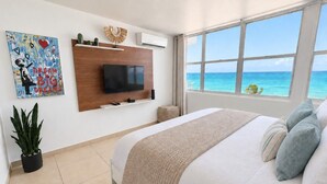 Een slaapkamer met een groot bed, een flatscreen televisie bevestigd aan een houten paneel, een potplant en uitzicht op de oceaan door grote ramen.