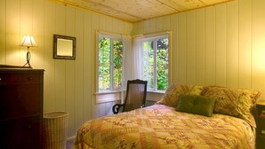Un dormitorio con techo de madera, una cama con colcha estampada, dos ventanas con cortinas, una silla, una lámpara y una pequeña cesta de mimbre.