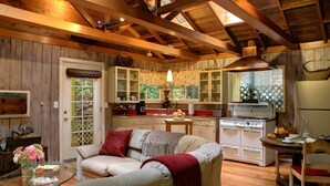 Una sala de estar acogedora con un sofá, una mesa de centro de vidrio y una zona de comedor con mesa y sillas. Se ve una cocina con armarios de madera y una estufa al fondo.