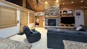 A spacious living room with a stone fireplace, wooden ceiling, and a large TV.