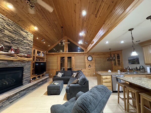 A cozy cabin interior with a stone fireplace, a large gray sectional sofa, a flat-screen TV, wooden ceiling, and a kitchen area with a granite countertop. - Luxury Knotty Pine Cabin IN THE Pinetop Lakes C.C (Pinetop-Lakeside)