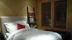 A neatly made bed with white bedding and a red pillow, a bedside table with a lamp, and a window with blinds.