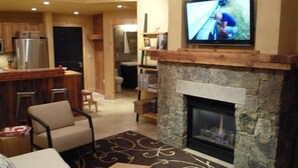 A living room with a stone fireplace, a television mounted above it, and a kitchen area in the background.