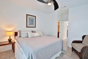 Une chambre à coucher comprenant un lit, une table de chevet, une lampe, un ventilateur de plafond, un tableau encadré accroché au mur et un fauteuil en osier.