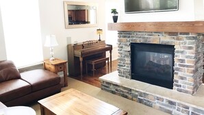 A living room with a stone fireplace, a television mounted above it, a piano, and a brown leather sofa.