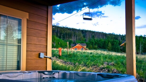 A wooden cabin with a hot tub and a view of a forest and a house in the distance.