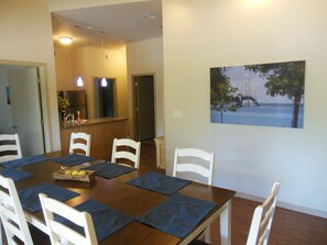 A dining area with a wooden table set for six, white chairs, and blue placemats. A kitchen is visible in the background, and a painting of a bridge over water hangs on the wall.