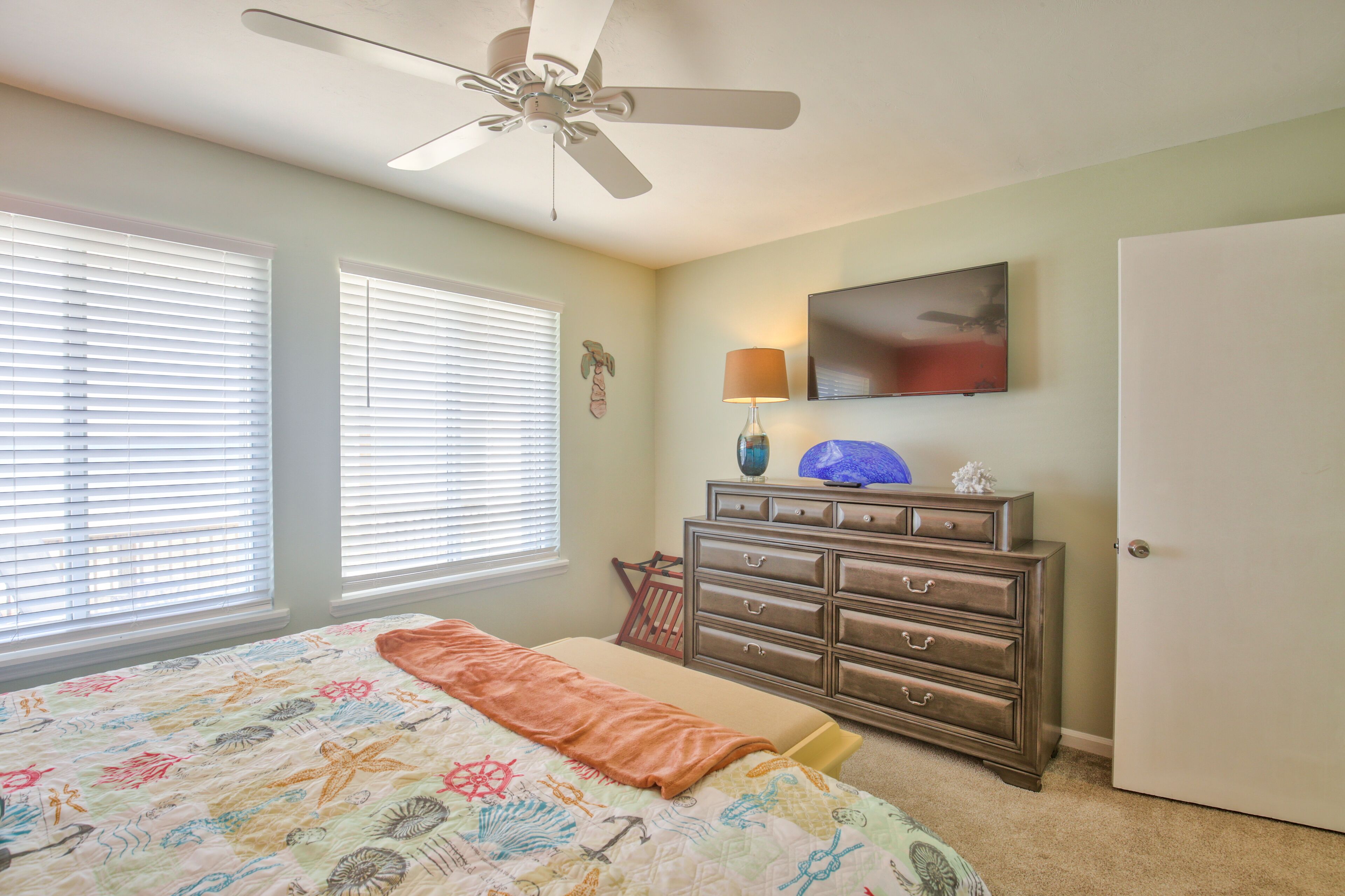A bedroom with a bed, dresser, ceiling fan, window blinds, and a television.