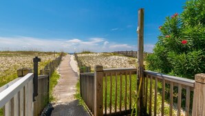 A wooden boardwalk leading to a beach with a wooden fence on one side and a bush with red flowers on the other.