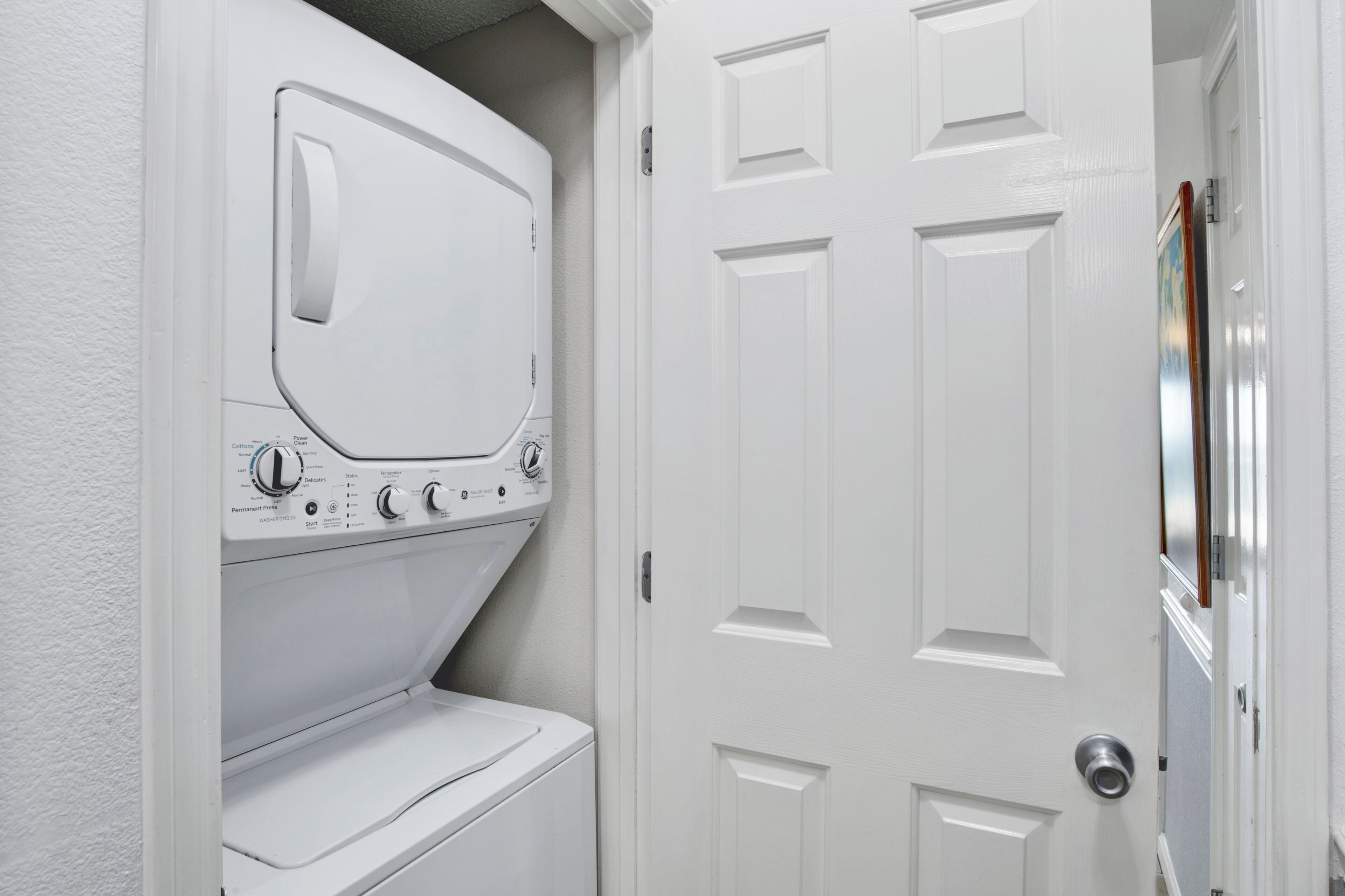 A laundry room with a stacked washer and dryer, a white door, and a framed picture on the wall.