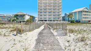 A beachfront hotel with a central building, a wooden boardwalk leading to it, and a sandy beach area.