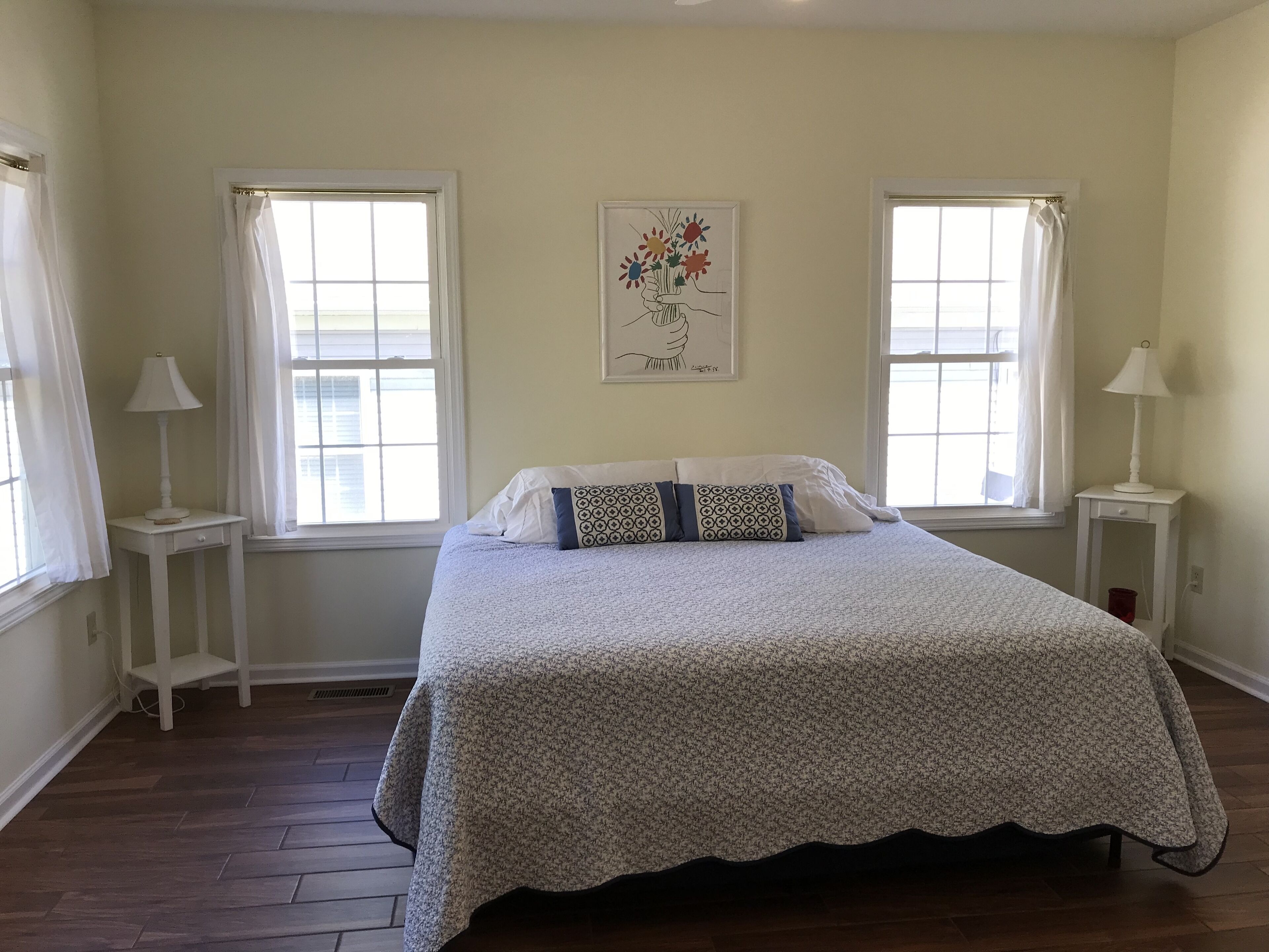 A bedroom with a bed, two windows, a nightstand with a lamp, and a wall-mounted artwork.