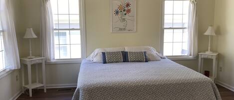 A bedroom with a bed, two windows, a nightstand with a lamp, and a wall-mounted artwork.