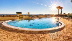 Une piscine circulaire à l’eau d’un bleu limpide, entourée d’une terrasse carrelée, et offrant une vue sur l’océan et les palmiers en arrière-plan.