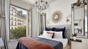 Una camera da letto con un letto grande, un lampadario, uno specchio a forma di sole e vista sugli edifici attraverso la finestra.