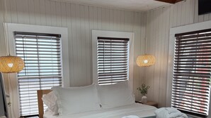 Un lit bien fait, avec du linge de lit blanc et un plaid turquoise, encadré par deux fenêtres équipées de stores, une lampe de chevet et une petite table de chevet sur laquelle se trouve un panier.