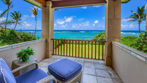 A covered balcony with a view of the ocean, featuring a wooden railing, a small table with a plant, and two cushioned chairs.