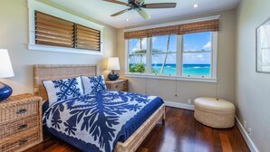 A bedroom with a bed, a ceiling fan, a window with a view of the ocean, and a wicker nightstand.