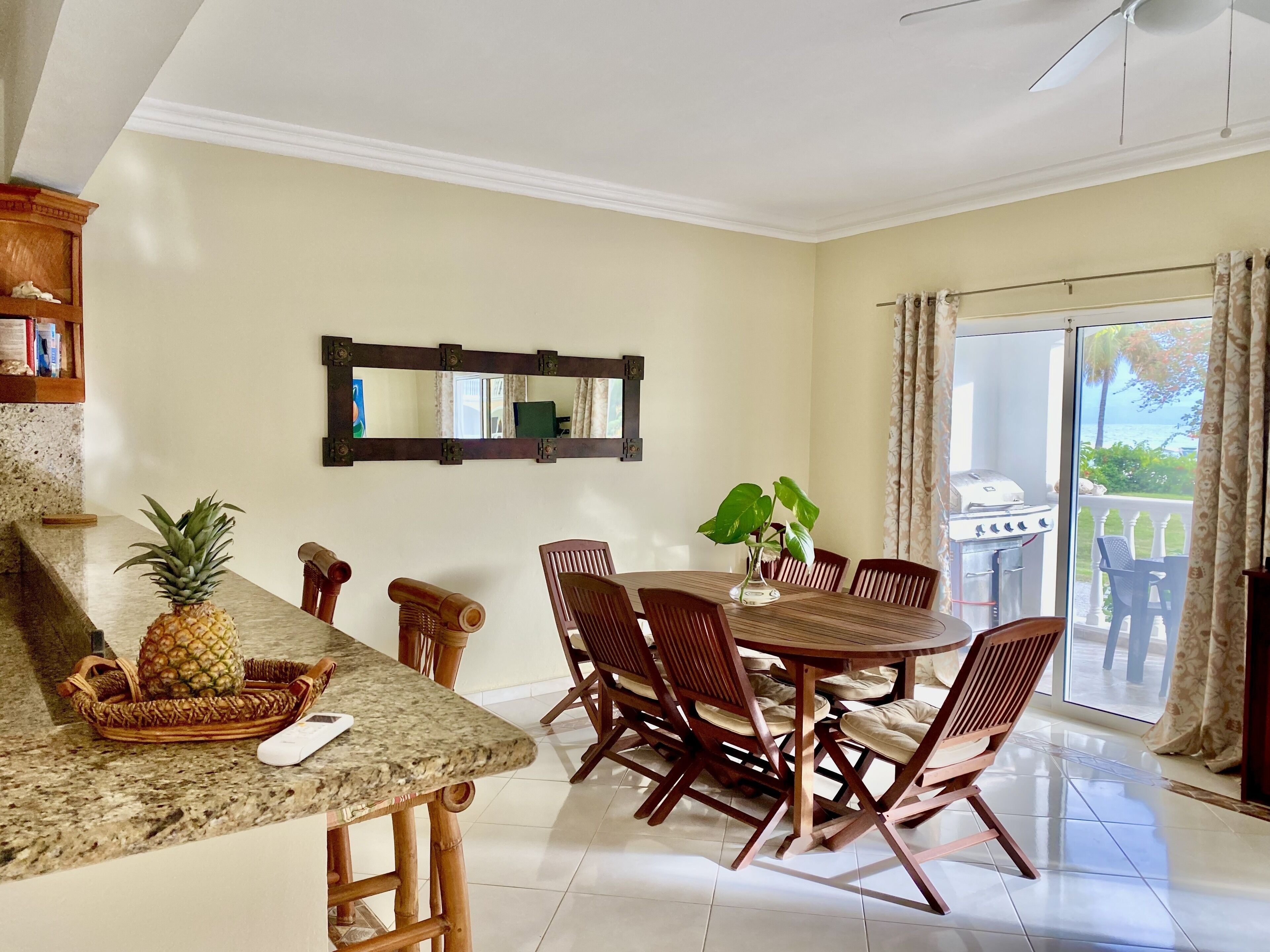A dining area with a round table and chairs, a kitchen counter with a pineapple, and a view of the outdoors through a sliding glass door.