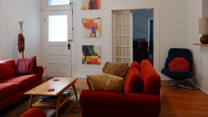 A living room with a red sofa, a wooden coffee table, and a blue armchair. There are abstract paintings on the wall and a door leading to another room.