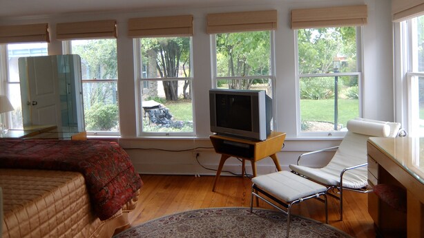 A room with a bed, a chair, a television on a wooden stand, and large windows with a view of greenery outside.