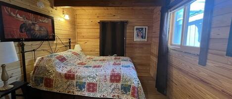 Ein gemütliches Blockhauszimmer mit Holzvertäfelung, einem Bett mit farbenfroher Quilttdecke, einer Lampe und einem Bild eines Elchs an der Wand.