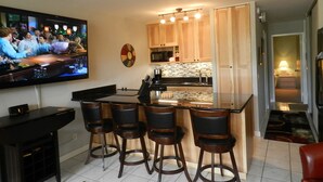 Un espacio habitable compacto con cocina, zona de comedor con taburetes de bar, televisor montado en la pared y una puerta visible que conduce a otra habitación.