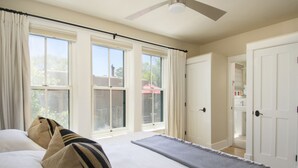 A bedroom with a large bed, a ceiling fan, and a view of the outdoors through large windows.