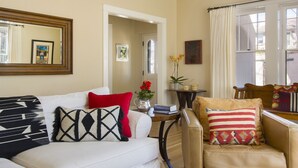 A living room with a white sofa, a red pillow, a patterned pillow, a small table with a vase of flowers, and a window with curtains.