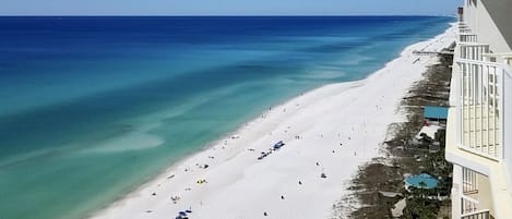 A beach with white sand and clear blue water. There are several umbrellas and people on the beach. A building with balconies overlooks the beach.