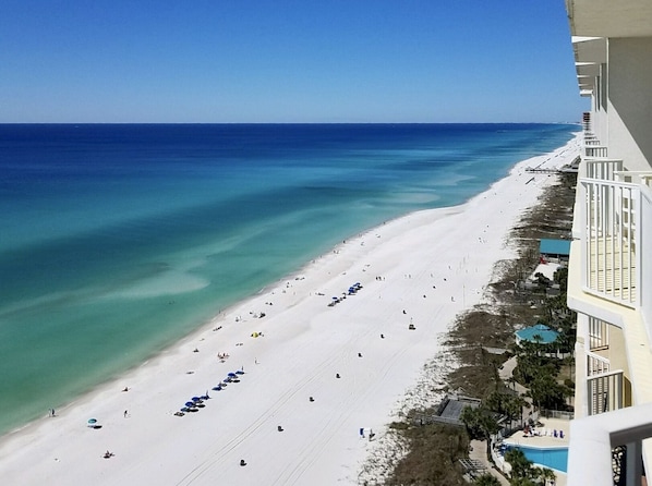 Una playa de arena blanca y agua azul cristalina. Varias sombrillas y gente hay en la playa. Un edificio con balcones domina la playa.