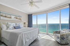 A bedroom with a large bed, a ceiling fan, a view of the ocean, and a balcony with a chair.