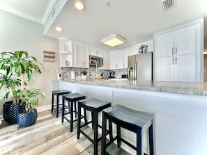 Cocina moderna con armarios blancos, refrigerador de acero inoxidable y encimera de mármol. Hay tres taburetes negros y dos plantas en macetas.