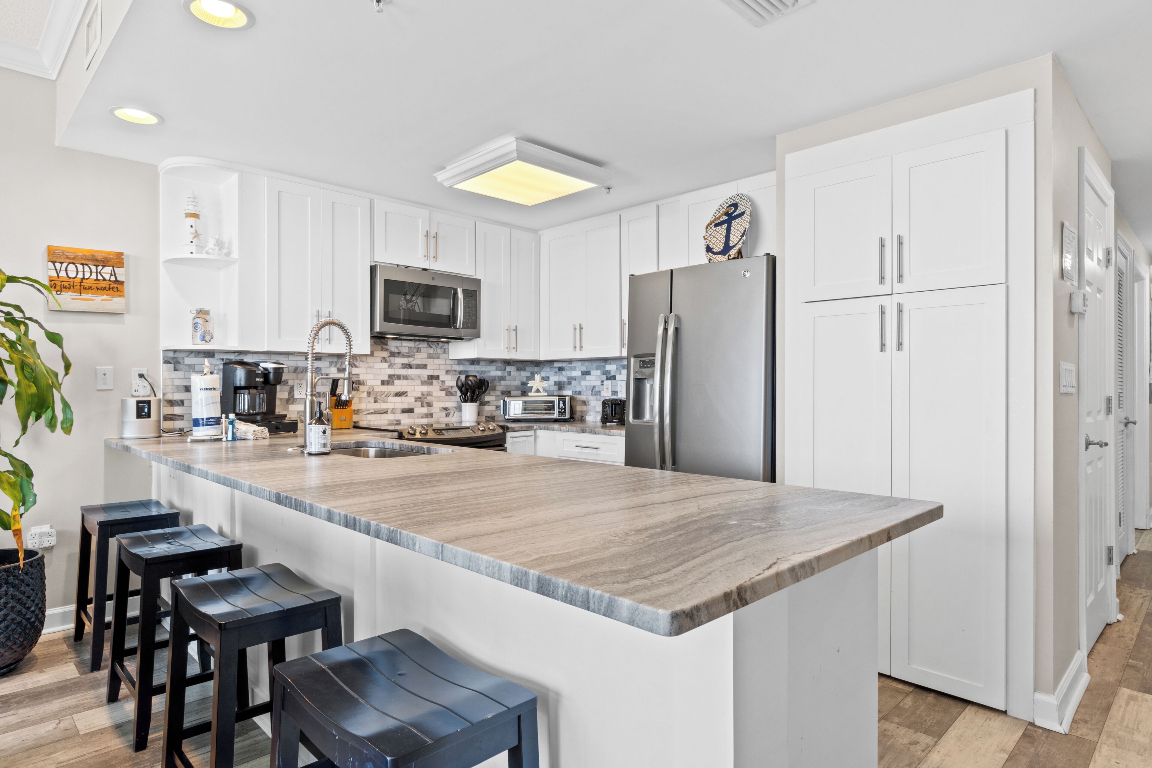 A modern kitchen with white cabinetry, a large island with bar stools, a stainless steel refrigerator, and a microwave.