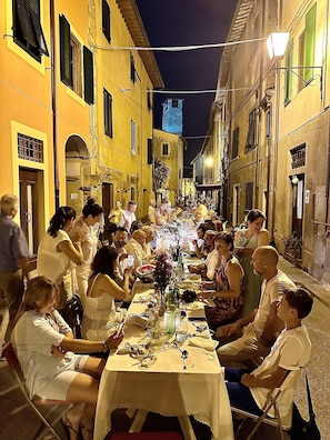 A group of people dining outdoors at a long table in a narrow street with buildings on either side.