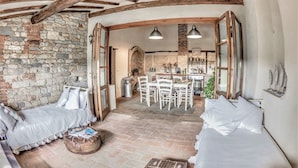 A rustic interior with exposed wooden beams, stone walls, a stone fireplace, and a dining area with a kitchen in the background.
