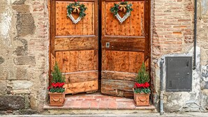 A rustic wooden double door with a heart-shaped wreath, flanked by potted plants with red flowers, set in a stone building with a brick archway.