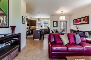 A spacious living room with a red leather sofa, a coffee table, and a TV stand. There is a kitchen in the background with modern appliances and a dining area with a table and chairs. - Urban Oasis in Downtown Walla Walla.  Walk to Wine, Restaurants and More! (Walla Walla)