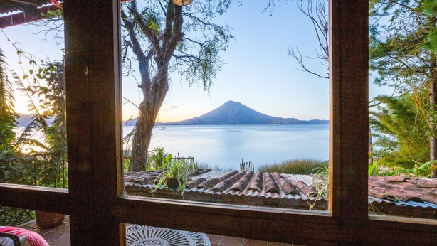 On aperçoit, à travers une fenêtre, un patio doté d’un toit en tuiles, d’une table et de chaises, d’où l’on a vue sur une étendue d’eau calme et une montagne.