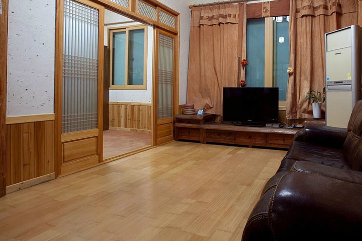 A traditional Japanese-style living room with a wooden floor, a brown leather sofa, a television, and a sliding door leading to another room.