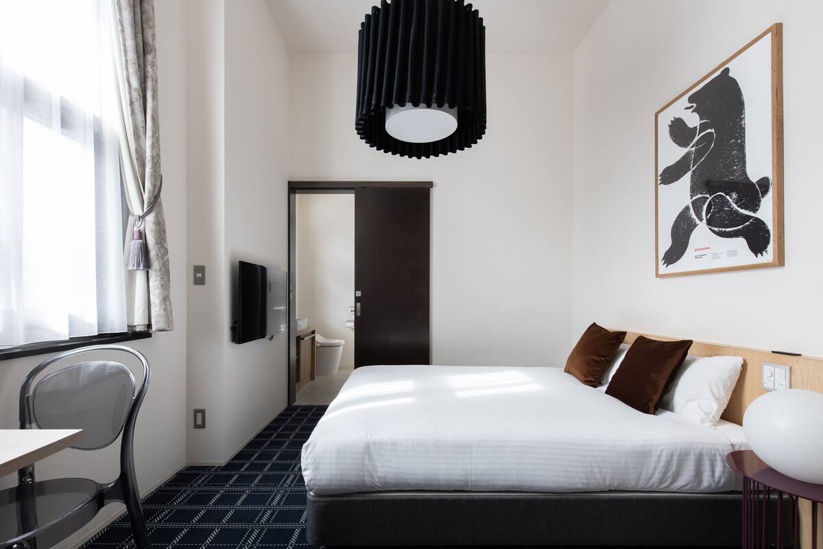 A hotel room with a bed, a desk, a chair, a TV, and a painting on the wall.