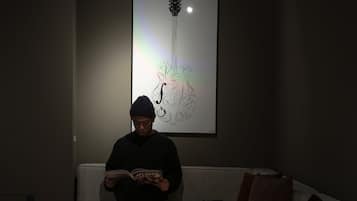 Une personne est assise sur un canapé, lisant un livre dans une pièce à la lumière tamisée, au décor moderne, avec une petite table et une œuvre d’art accrochée au mur.