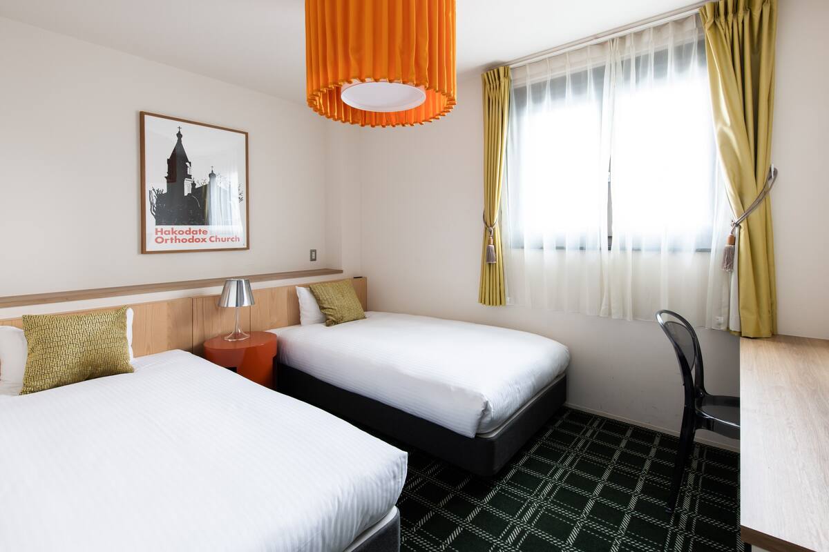 A hotel room with two beds, a desk, a chair, a hanging orange pendant light, a framed picture of a church, and a window with yellow curtains.