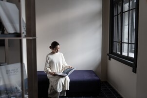 Una persona sentada en un banco leyendo un libro en una habitación con ventana y estantería.