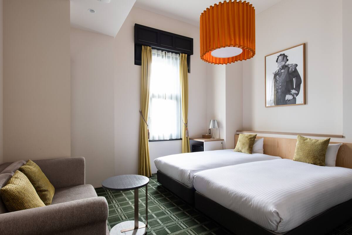 A hotel room with a bed, a chair, a small table, a lamp, and a painting on the wall.